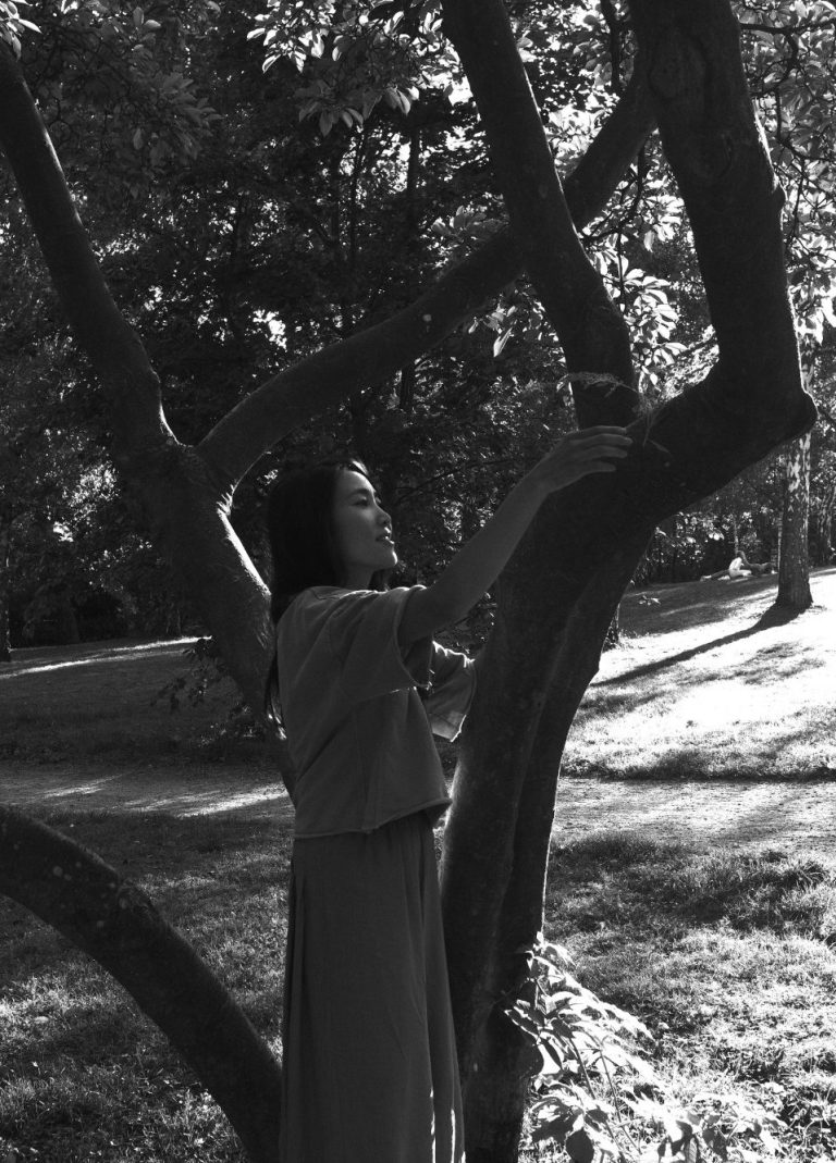 Eine Frau mit langen schwarzen Haaren und weiter Kleidung mit asiatischen Wurzeln umgreift einen Ast von einem Baum. Sie befinden sich in einem Park, die Sonne scheint. Das Foto ist in schwarz weiß.